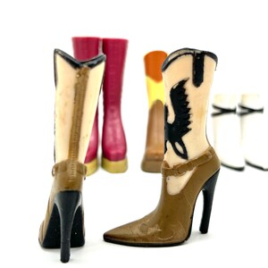 May include: A collection of miniature cowboy boots in various colors, including brown, tan, and red. The boots have high heels and decorative details, with one featuring a black bird design. The boots are set against a white background.
