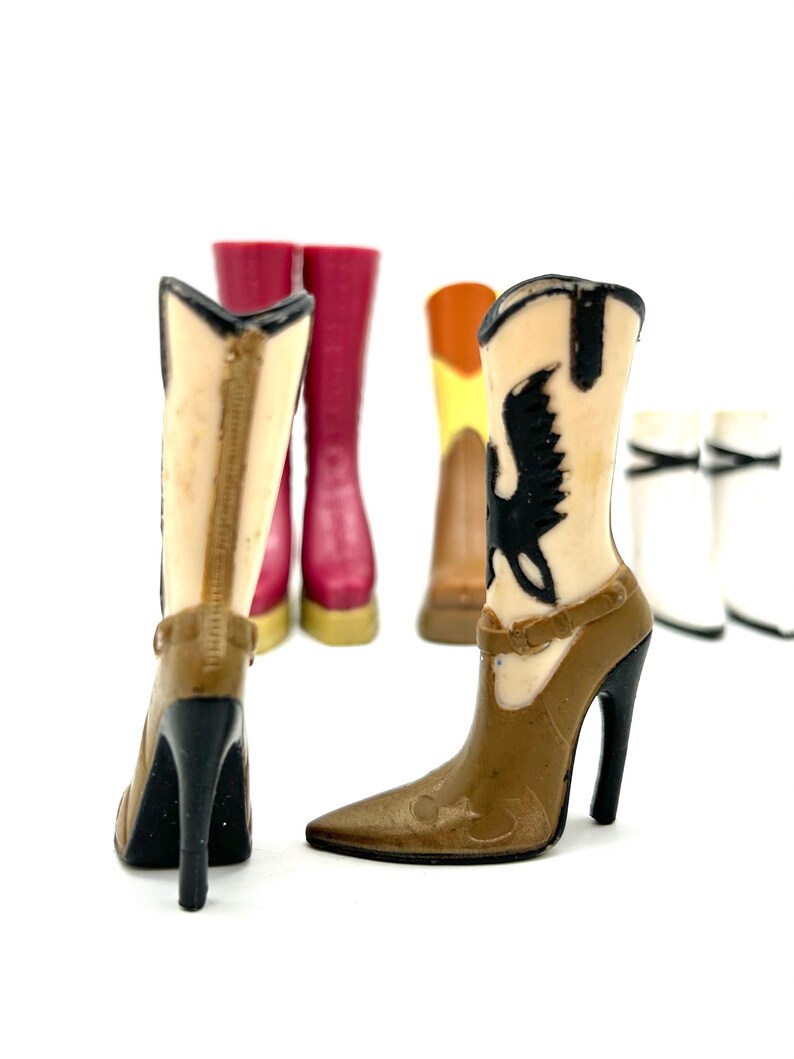 May include: A collection of miniature cowboy boots in various colors, including brown, tan, and red. The boots have high heels and decorative details, with one featuring a black bird design. The boots are set against a white background.