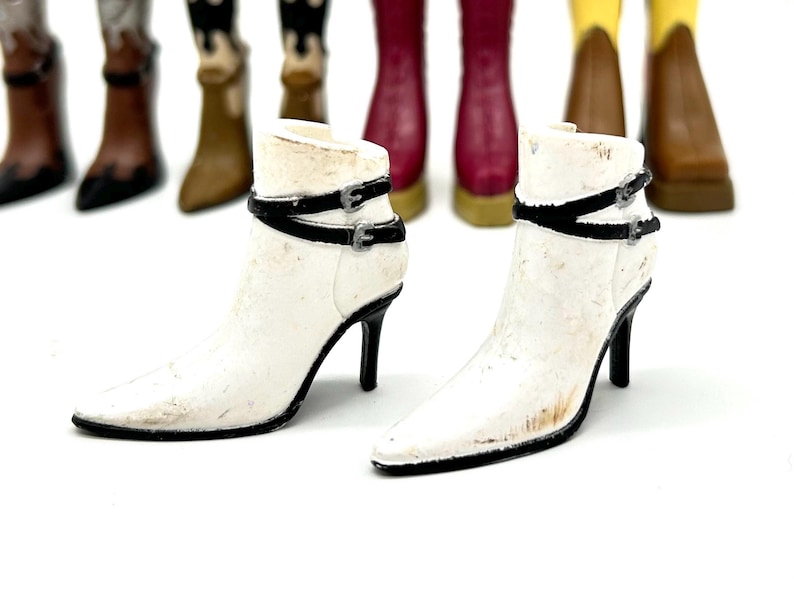 May include: Two white ankle boots with black straps and buckles, set against a backdrop of various colored cowboy boots. The boots have pointed toes and stiletto heels. The white boots are the focal point.