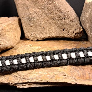 May include: Black and white paracord bracelet with a black plastic buckle. The bracelet is made with reflective white paracord.