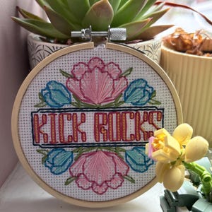 Può includere: Un telaio da ricamo a punto croce con le parole "KICK ROCKS" in uno stendardo rettangolare. Il disegno presenta fiori rosa e turchese. Il telaio è ambientato su uno sfondo con una pianta succulenta in un vaso decorativo.