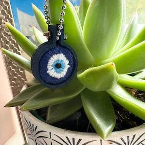 May include: A blue and white evil eye charm with a silver chain attached. The charm is hanging from a succulent plant in a white pot with a black and white pattern.