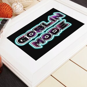 Op de afbeelding: Een wit ingelijst print met een zwarte achtergrond en de tekst "GORLIN MODE" in neonpaarse en blauwe letters. De achtergrond is een galaxie-achtig patroon met paarse en blauwe kleuren.