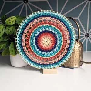 May include: A colorful crocheted mandala wall hanging with a blue, green, orange, and red color scheme. The mandala is mounted on a gold metal stand.