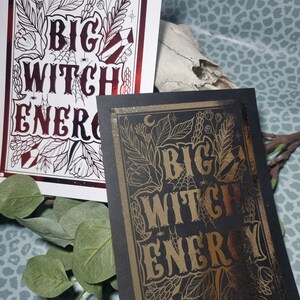 Op de afbeelding: Twee prints met de tekst "Big Witch Energy" in een gotisch lettertype. De print aan de linkerkant is op wit papier met rode en zwarte inkt. De print aan de rechterkant is op zwart papier met gouden inkt.