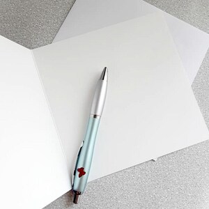 May include: A light blue pen with silver accents and a red floral design rests on a white greeting card. The pen is positioned near the center of the card, with a second card visible in the background. The background is a speckled gray surface.