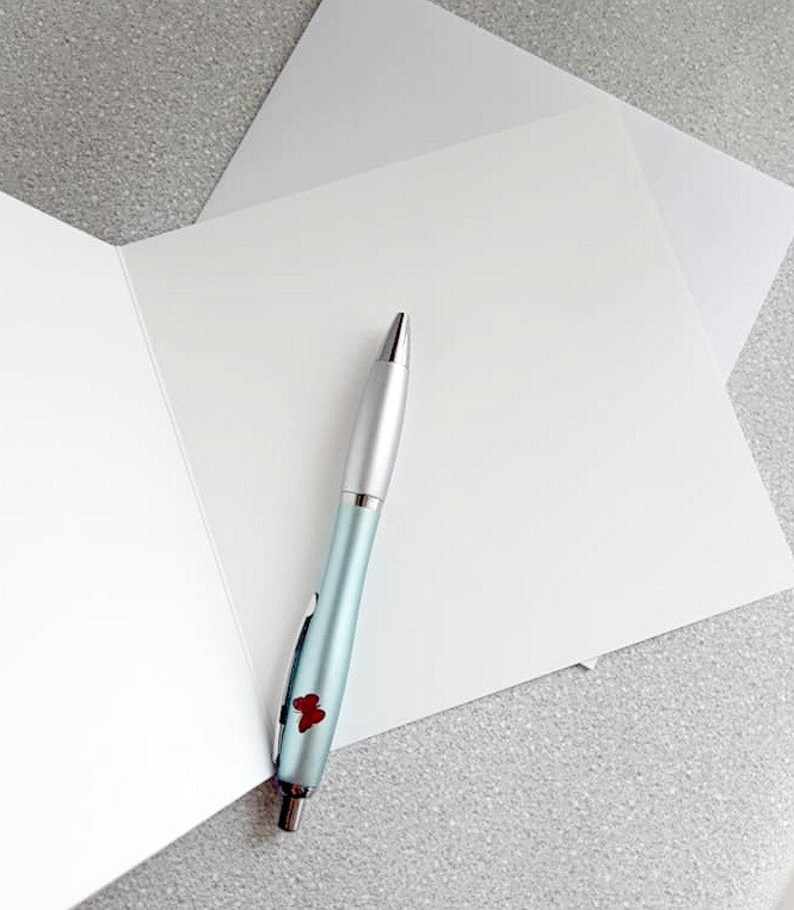 May include: A light blue pen with silver accents and a red floral design rests on a white greeting card. The pen is positioned near the center of the card, with a second card visible in the background. The background is a speckled gray surface.