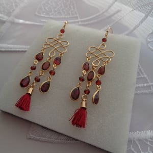 Pu&ograve; includere: Un paio di orecchini dorati con gocce di pietre preziose rosse e nappe rosse. Gli orecchini presentano un design delicato e intricato.