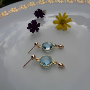 Könnte beinhalten: Ein Paar goldene Ohrringe mit hellblauen Edelstein-Akzenten. Die Ohrringe haben einen runden Edelstein mit einer goldenen Fassung und eine kleine goldene Perle oben.