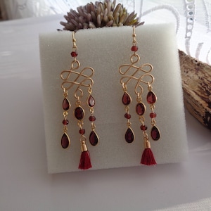 Pu&ograve; includere: Orecchini pendenti dorati con un designo di pietre preziose rosse. Gli orecchini presentano un design annodato con pi&ugrave; pietre preziose rosse e un fiocco.
