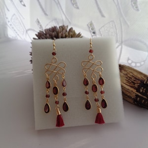 Pu&ograve; includere: Orecchini dorati con nappa rossa e gocce di pietre preziose rosse. Gli orecchini presentano un design delicato e intrecciato.