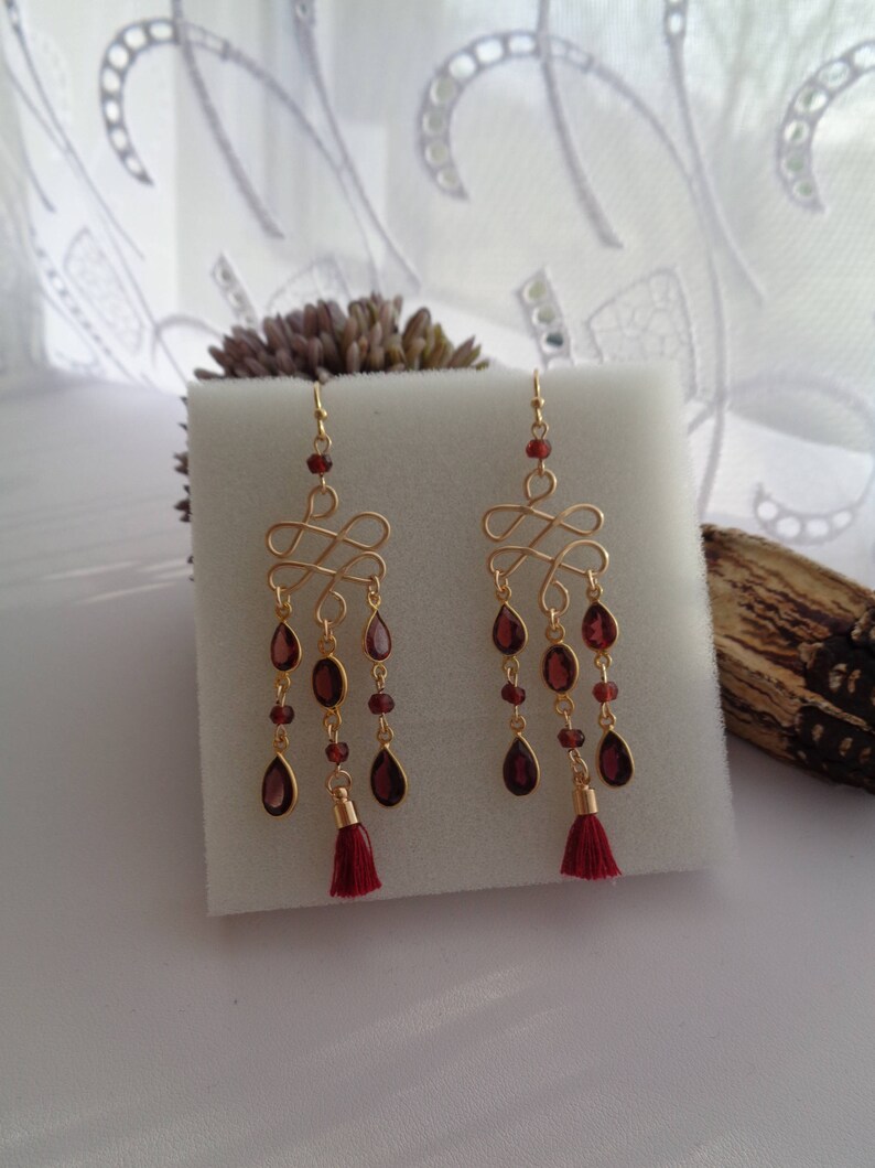 Pu&ograve; includere: Orecchini dorati con nappa rossa e gocce di pietre preziose rosse. Gli orecchini presentano un design delicato e intrecciato.