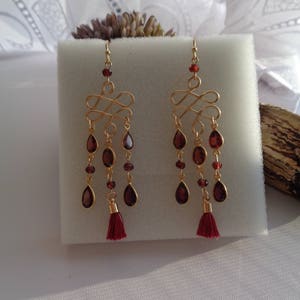 Pu&ograve; includere: Un paio di orecchini pendenti in oro con gocce di pietre preziose rosse e nappe rosse. Gli orecchini presentano un design delicato e intrecciato.