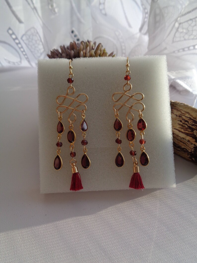 Pu&ograve; includere: Un paio di orecchini pendenti in oro con gocce di pietre preziose rosse e nappe rosse. Gli orecchini presentano un design delicato e intrecciato.