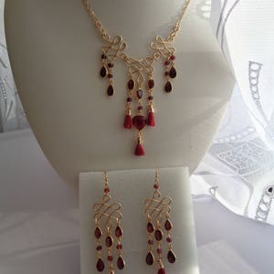 Pu&ograve; includere: Un set di collana e orecchini in oro con un ciondolo di pietra rossa e dettagli di nappe rosse. La collana presenta un design delicato e intrecciato, e gli orecchini hanno un design simile con pietre rosse pendenti.