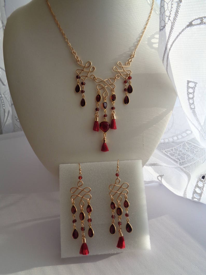 Pu&ograve; includere: Un set di collana e orecchini in oro con un ciondolo di pietra rossa e dettagli di nappe rosse. La collana presenta un design delicato e intrecciato, e gli orecchini hanno un design simile con pietre rosse pendenti.