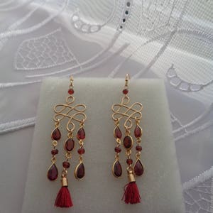 Pu&ograve; includere: Orecchini dorati con un design a goccia con gocce di pietre preziose rosse e nappe rosse.