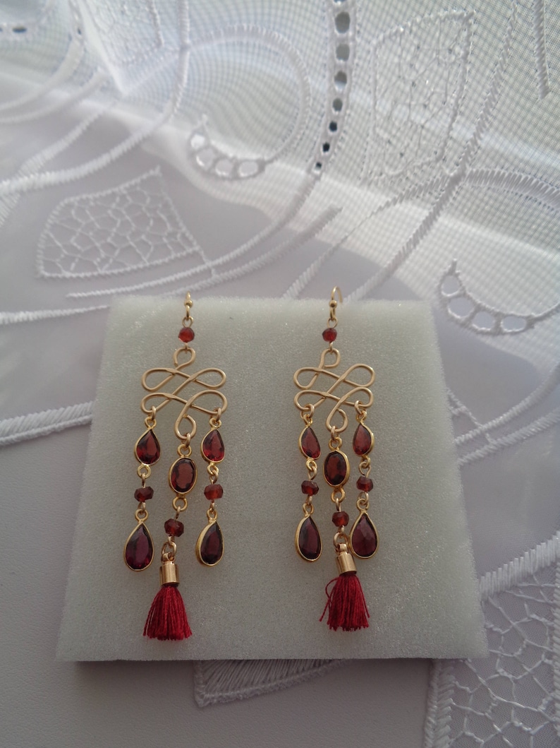 Pu&ograve; includere: Orecchini dorati con un design a goccia con gocce di pietre preziose rosse e nappe rosse.
