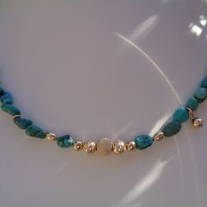 Op de afbeelding: Een turquoise en gouden ketting met een getextureerde gouden kraal in het midden. De ketting is gemaakt van kleine, onregelmatige turquoise stenen.