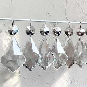 May include: A row of clear crystal chandelier pendants hanging from a white wire. Each pendant has a faceted, teardrop shape with a circular top and a metal hook for hanging. The background is a textured, off-white wall.