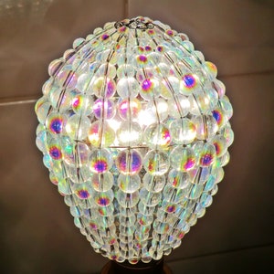 May include: A clear glass light bulb covered in iridescent beads, creating a shimmering effect. The beads are arranged in a spherical shape, with a wire frame holding them together. The light bulb is lit, casting a warm glow.