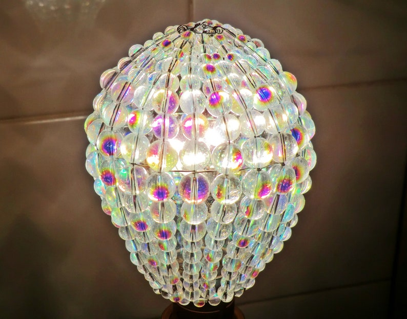 May include: A clear glass light bulb covered in iridescent beads, creating a shimmering effect. The beads are arranged in a spherical shape, with a wire frame holding them together. The light bulb is lit, casting a warm glow.
