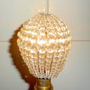 May include: A clear glass lampshade made of small, round beads. The beads are arranged in a spherical shape and are held together by a metal frame. The lampshade is lit from within, creating a warm, inviting glow.