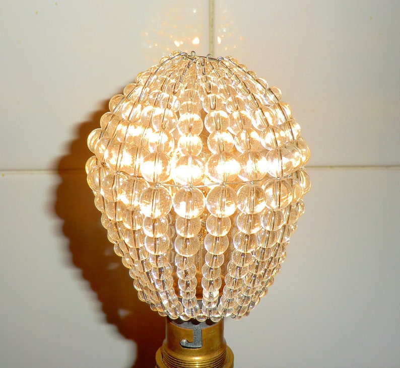 May include: A clear glass lampshade made of small, round beads. The beads are arranged in a spherical shape and are held together by a metal frame. The lampshade is lit from within, creating a warm, inviting glow.