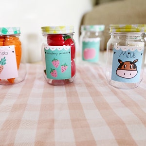 May include: Four glass jars with colorful lids and labels. The labels have hand-drawn illustrations and text. The jars contain carrots, strawberry preserves, an apple, and milk.