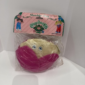 May include: A Cabbage Patch Kids shoulder bag in original plastic packaging. The bag is shaped like a cabbage with a beige face and magenta leaves. The header card features the Cabbage Patch Kids logo, illustrations of two children, and text.