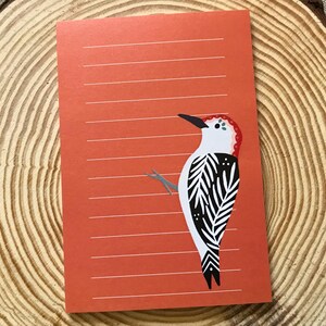 May include: A lined orange notepad featuring a stylized woodpecker illustration. The bird has a black beak, red head, white chest, and black and white patterned wings. The notepad is placed on a wooden slice.