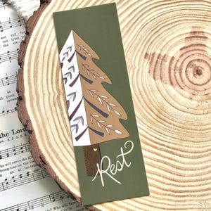 May include: A green bookmark with a stylized brown and white Christmas tree design. The word "Rest" is written in white script at the bottom. The bookmark is on a wooden slice with sheet music in the background.