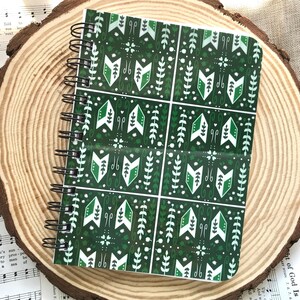 May include: A spiral-bound notebook featuring a repeating dark green and white pattern of stylized arrows and botanical elements. The cover has a grid layout with six rectangular sections, each containing a symmetrical design of white arrows.