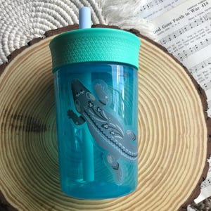 May include: A clear blue plastic tumbler with a teal lid and straw. A gray lizard sticker with floral accents decorates the side. The tumbler is designed for cold beverages.