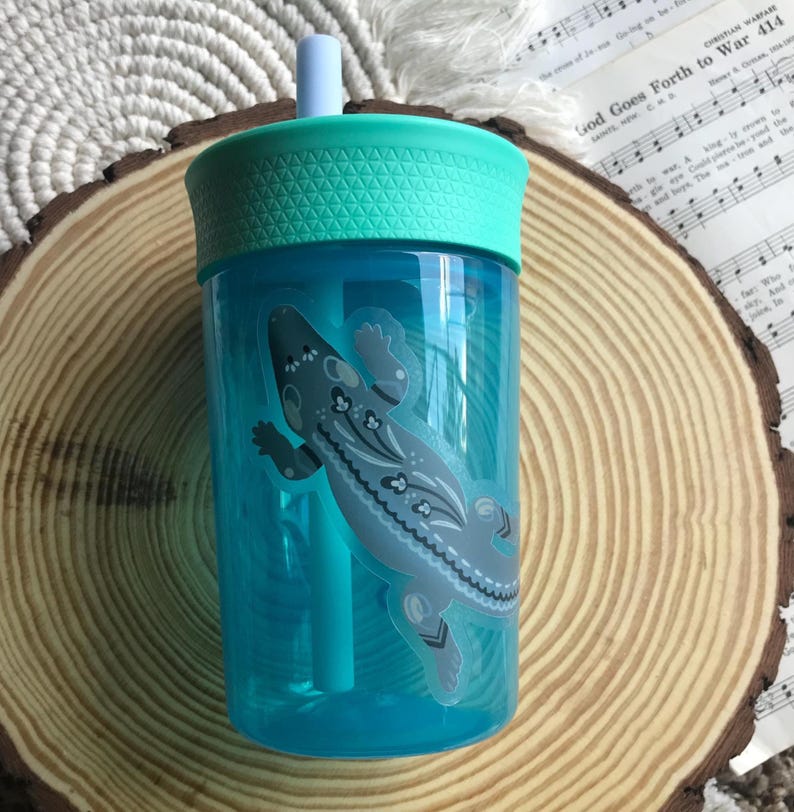 May include: A clear blue plastic tumbler with a teal lid and straw. A gray lizard sticker with floral accents decorates the side. The tumbler is designed for cold beverages.
