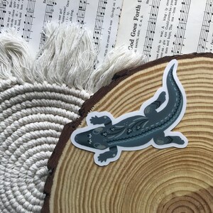 May include: A sticker featuring a stylized alligator in shades of blue and gray with decorative patterns. The sticker is placed on a wooden slice, with a macrame coaster and sheet music in the background.