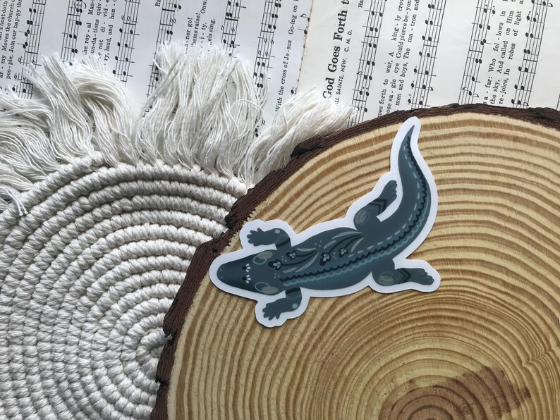 May include: A sticker featuring a stylized alligator in shades of blue and gray with decorative patterns. The sticker is placed on a wooden slice, with a macrame coaster and sheet music in the background.