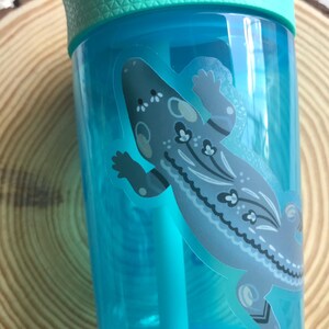May include: A clear, turquoise water bottle with a light green lid and a straw. The bottle features a decorative sticker of a gray and blue lizard with floral patterns. The bottle is partially visible against a wooden background.