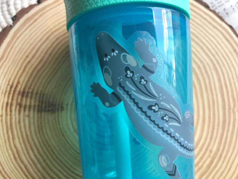 May include: A clear, turquoise water bottle with a light green lid and a straw. The bottle features a decorative sticker of a gray and blue lizard with floral patterns. The bottle is partially visible against a wooden background.