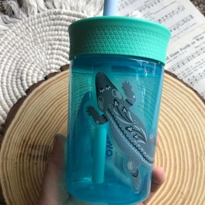 May include: A clear blue plastic tumbler with a teal lid and straw. The tumbler features a decorative sticker of a gray and white lizard. The tumbler is designed for cold beverages.