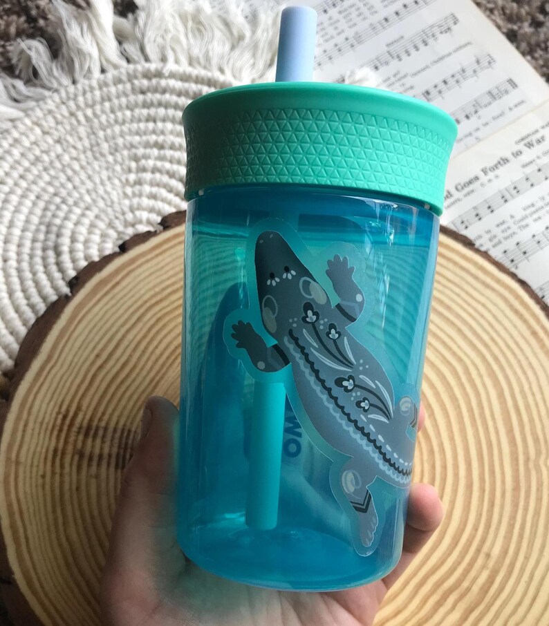 May include: A clear blue plastic tumbler with a teal lid and straw. The tumbler features a decorative sticker of a gray and white lizard. The tumbler is designed for cold beverages.