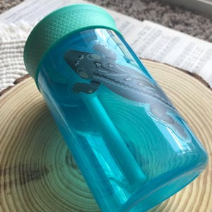 May include: A clear, turquoise water bottle with a teal lid and straw. The bottle features a stylized illustration of a gray and white salamander. The bottle is resting on a wooden slice.