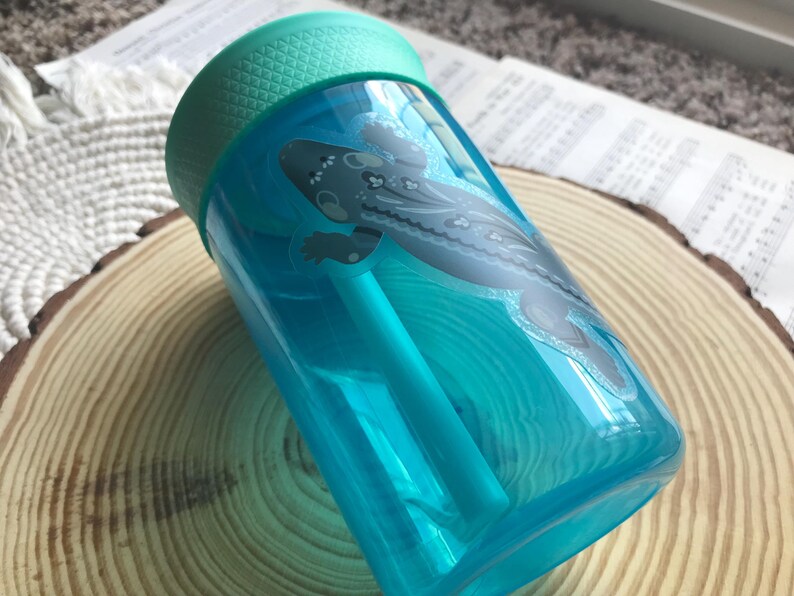 May include: A clear, turquoise water bottle with a teal lid and straw. The bottle features a stylized illustration of a gray and white salamander. The bottle is resting on a wooden slice.