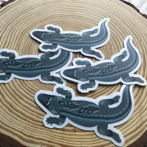 May include: Four stickers featuring a stylized lizard design in shades of gray and blue. The lizards have decorative patterns on their backs and are arranged on a wooden surface.