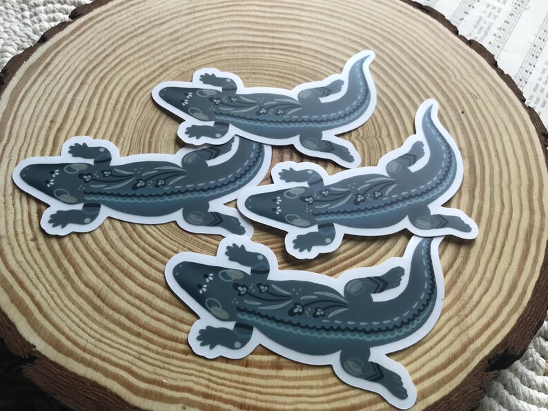 May include: Four stickers featuring a stylized lizard design in shades of gray and blue. The lizards have decorative patterns on their backs and are arranged on a wooden surface.