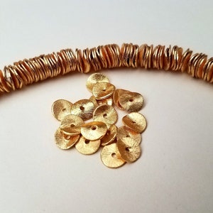 May include: A pile of gold-colored metal beads, each with a flat, round shape and a slightly textured surface. The beads are arranged in a loose pile, with some overlapping.