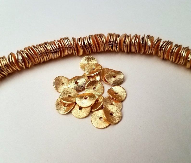 May include: A pile of gold-colored metal beads, each with a flat, round shape and a slightly textured surface. The beads are arranged in a loose pile, with some overlapping.