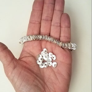 May include: A silver bracelet made of small, round, flat discs. The bracelet is shown on a hand, along with a pile of loose discs.