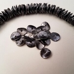 May include: A pile of black metal sequins with a slightly iridescent finish. The sequins are round with a small hole in the center.
