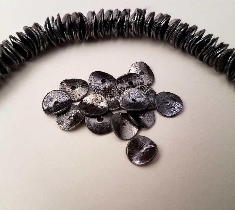 May include: A pile of black metal sequins with a slightly iridescent finish. The sequins are round with a small hole in the center.